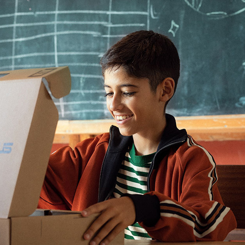 Een lachende student opent een ASUS-laptopdoos, zittend voor een schoolbord in een klaslokaal