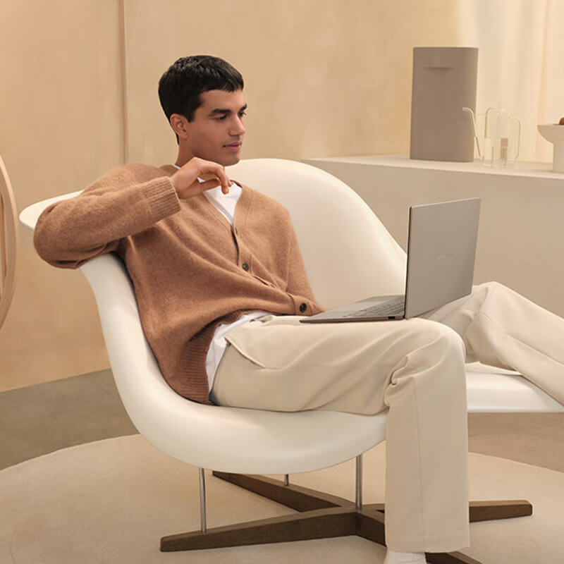 A man sits in a comfortable, laid-back position on a white designer armchair with an ASUS Zenbook A14 laptop on his lap, looking at its screen