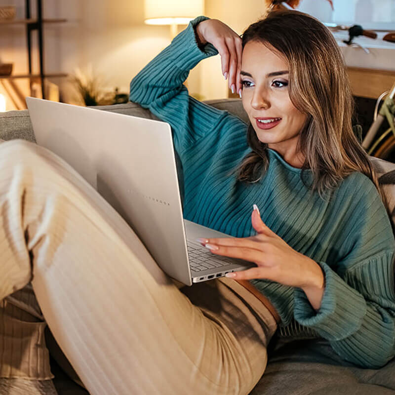 A young woman sits on the sofa while she uses an ASUS Vivobook S16 laptop