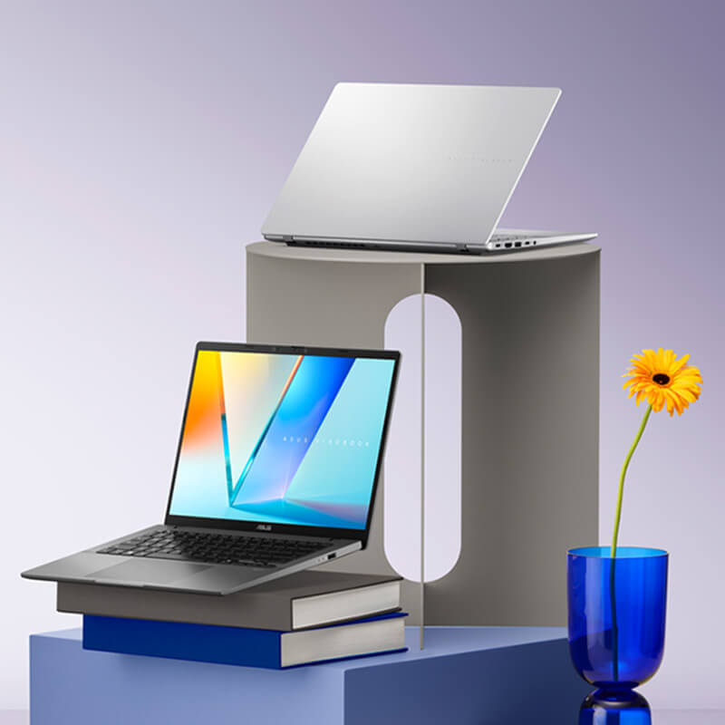 Two open ASUS Vivobook S14 laptops sit on two stands of different geometric shapes, accompanied by a vase with one flower in it. Both laptops sit at angles and are open, with one facing away and one facing forward.