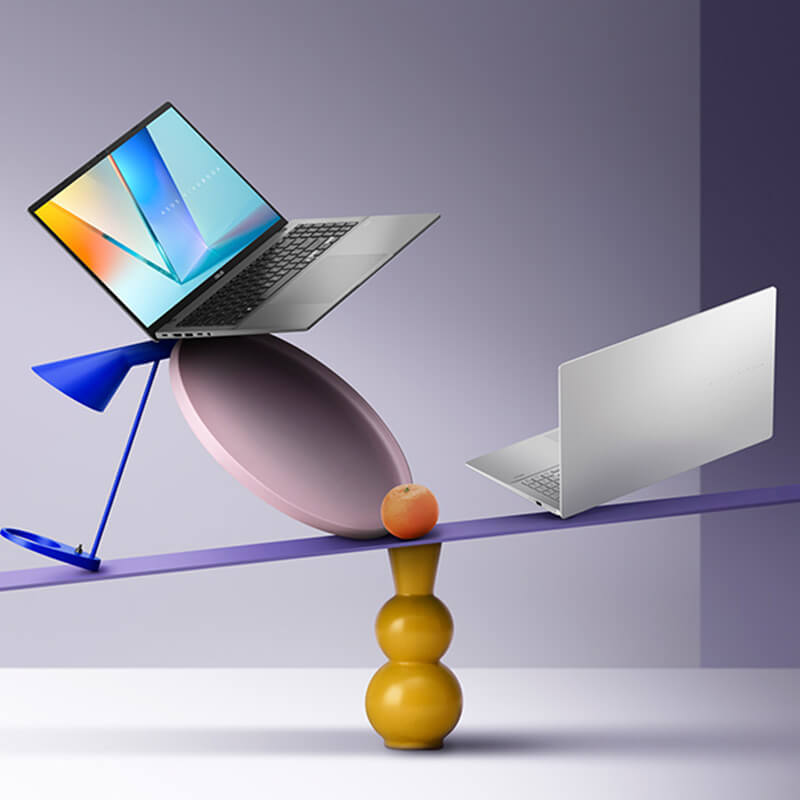 A Matte Gray ASUS Vivobook S16 is open while a Cool Silver ASUS Vivobook S16 is closed. Both laptops are balanced on a colorful, artistic seesaw with geometric shapes set against a soft purple gradient background.