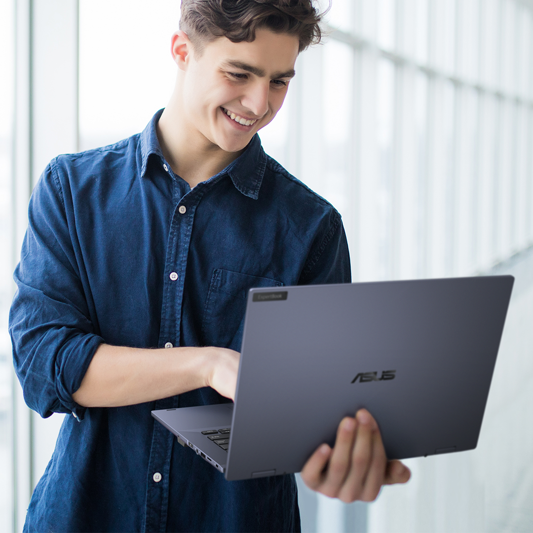 一名員工手持 ASUS ExpertBook 筆電，站在走廊上面帶微笑看著筆電。
