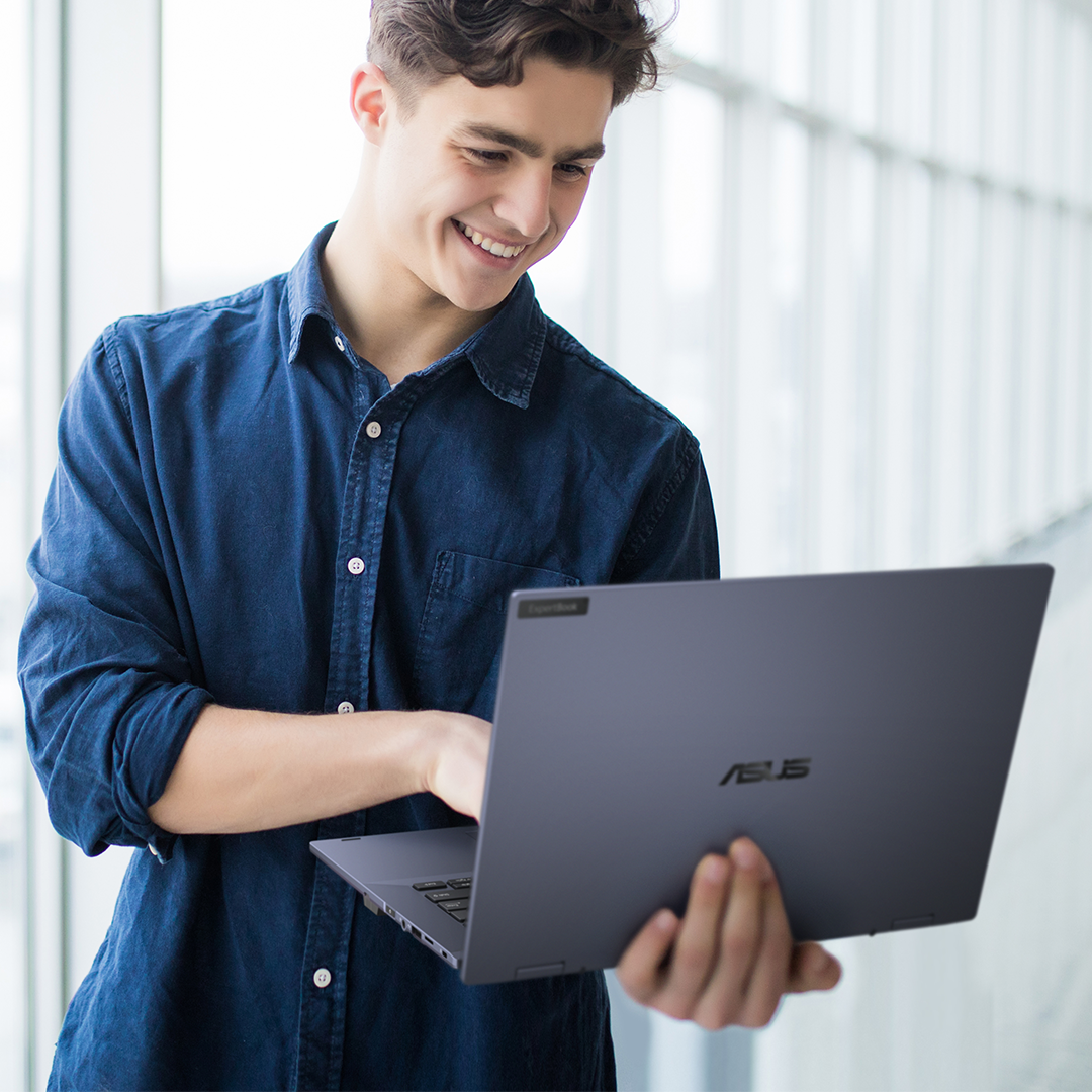 An employee holds ASUS ExpertBook on hand, looking on it with smile and standing at corridor.