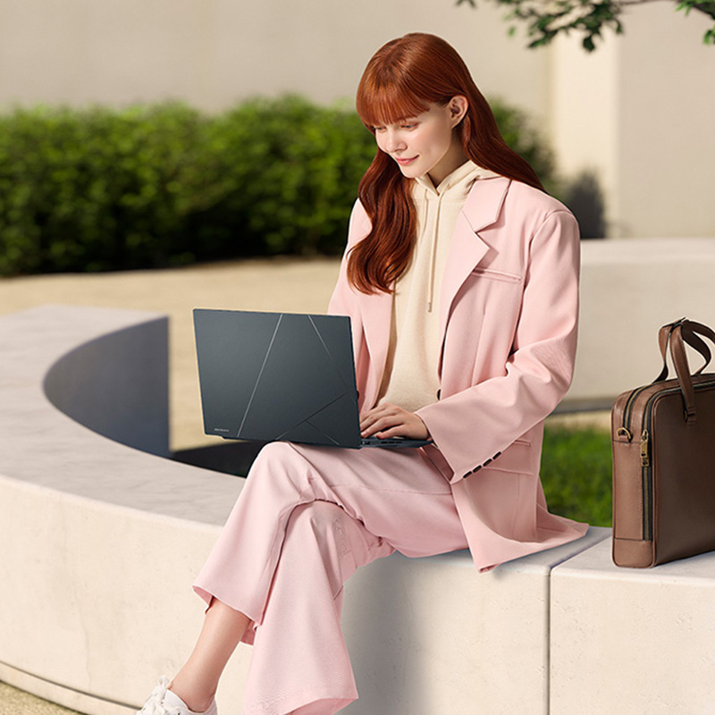 Девушка сидит на ограде с ноутбуком ASUS Zenbook 14X OLED на коленях. Рядом с ней стоит коричневый портфель, а на заднем фоне видно дерево