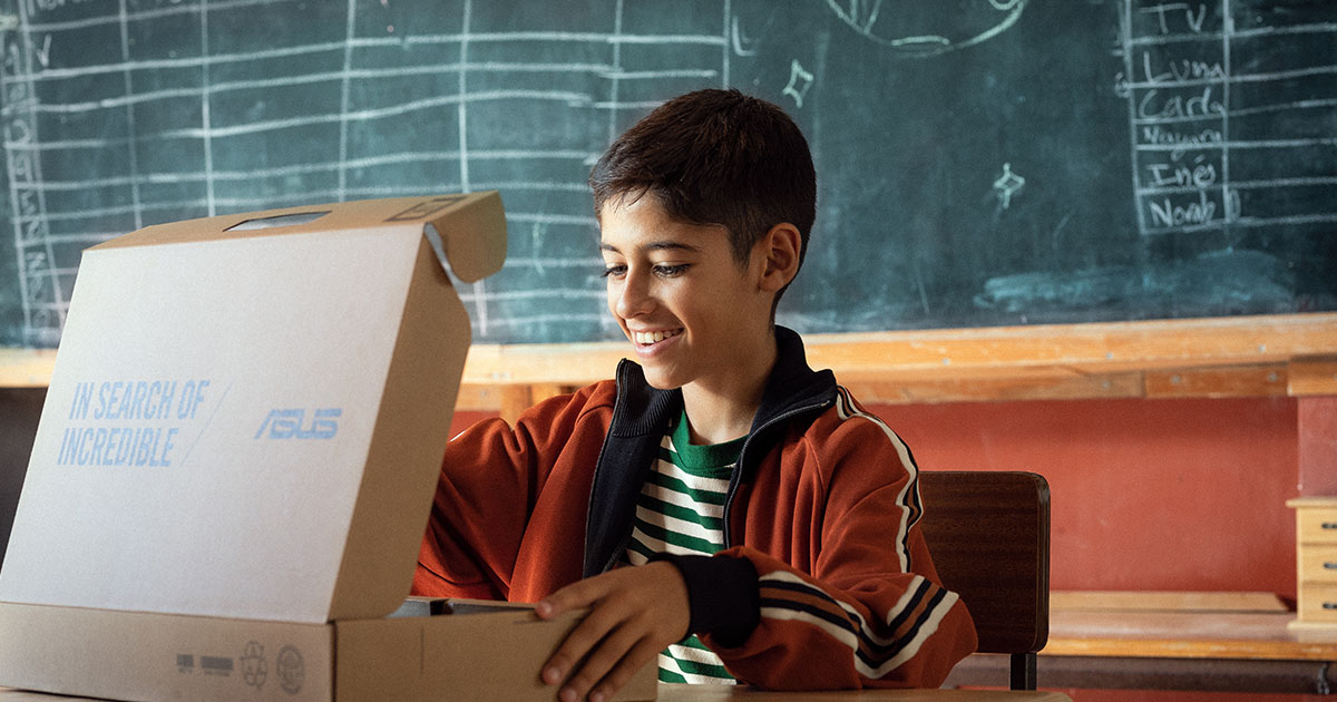 Een lachende student opent een ASUS-laptop, zittend voor een schoolbord in een klaslokaal