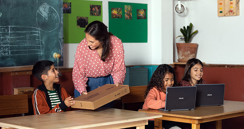 a teacher handing an ASUS laptop box to a smiling young male student in a classroom while two female students are looking at their ASUS laptop screens