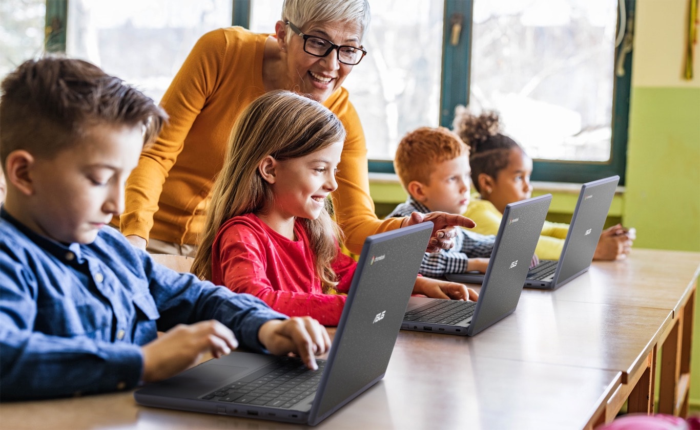 Three angled rear view of ASUS Chromebook CR11 in laptop mode on the desk. Three children are using them for the class with a female teacher standing in the back supporting them. Another girl in the last row is looking forward.