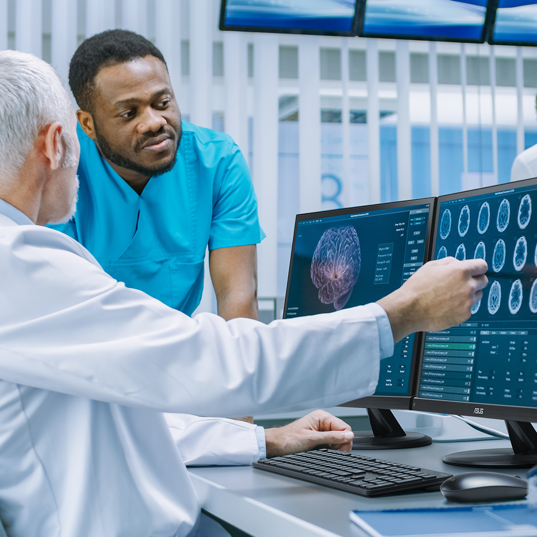 Due operatori sanitari guardano le immagini cerebrali proiettate su tre monitor ASUS e discutono tra loro.
