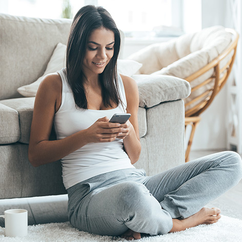 A woman at home is using a smartphone while leaning against a sofa, with a WiFi signal circle on the phone indicating a connection to the internet.