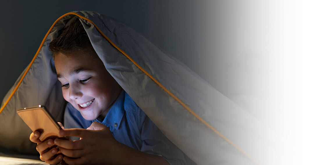 Een jongen verstopt zich onder een deken en gebruikt vrolijk een mobiele telefoon voor het slapengaan.