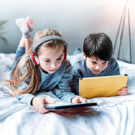 two children lying on a bed using their tablets.