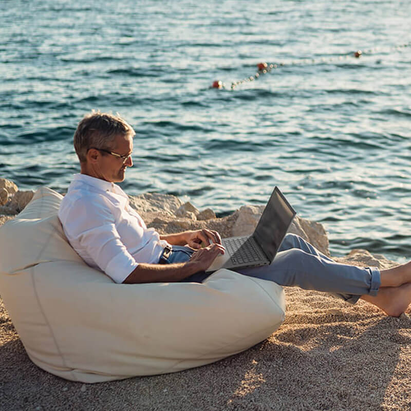a middle-aged man sitting on a beach on a bean bag with an ASUS Vivobook laptop on his lap