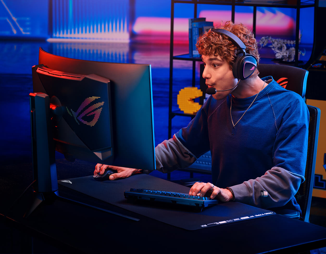 A man with curly hair sits at a gaming desk, wearing the ROG Delta II with a microphone. He is intensely focused on the ROG monitor with excitement, holding a ROG gaming mouse with one hand and a keyboard with the other.