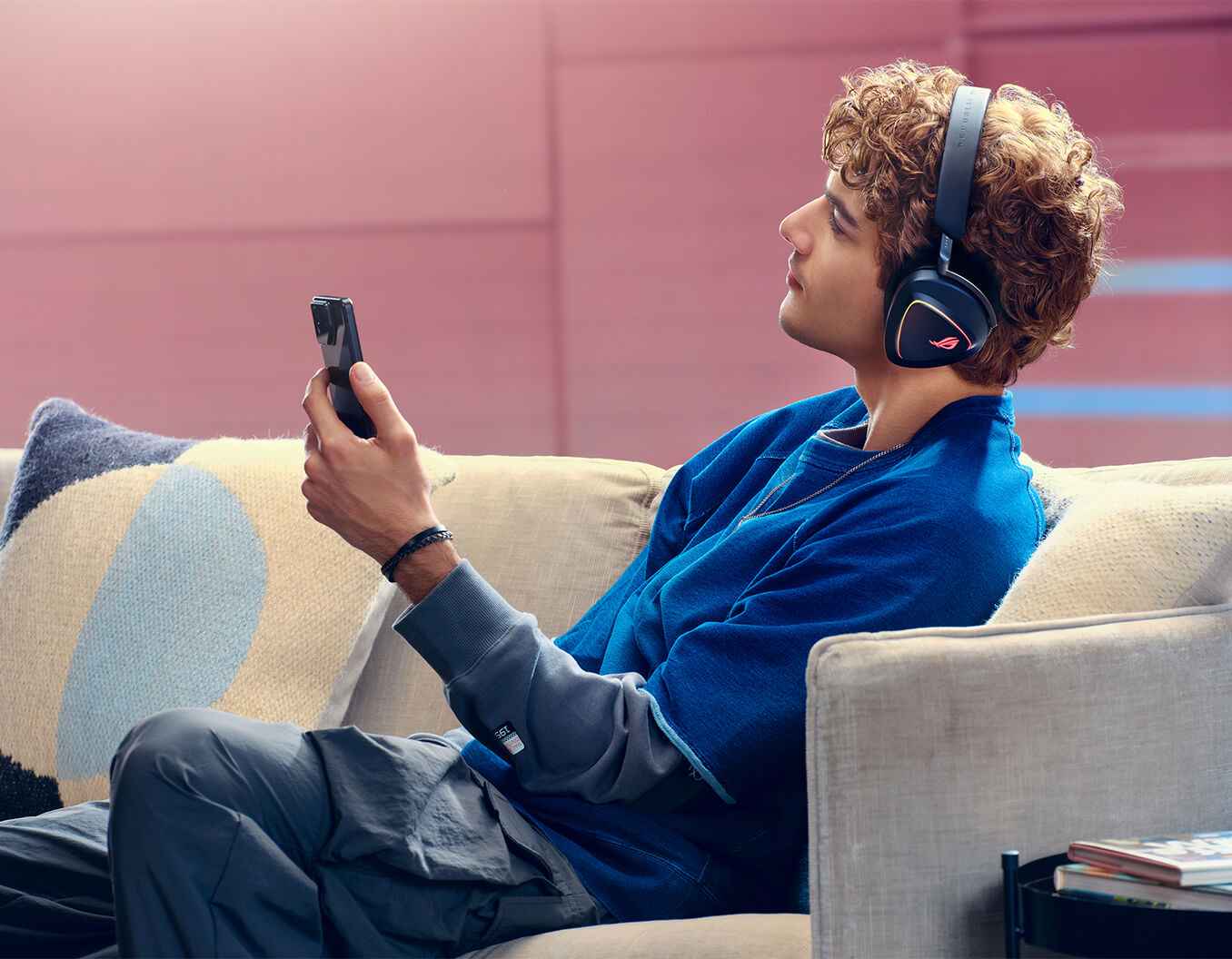 A man with curly hair wearing ROG Delta II sits on the sofa and holds a ROG phone on the left hand.