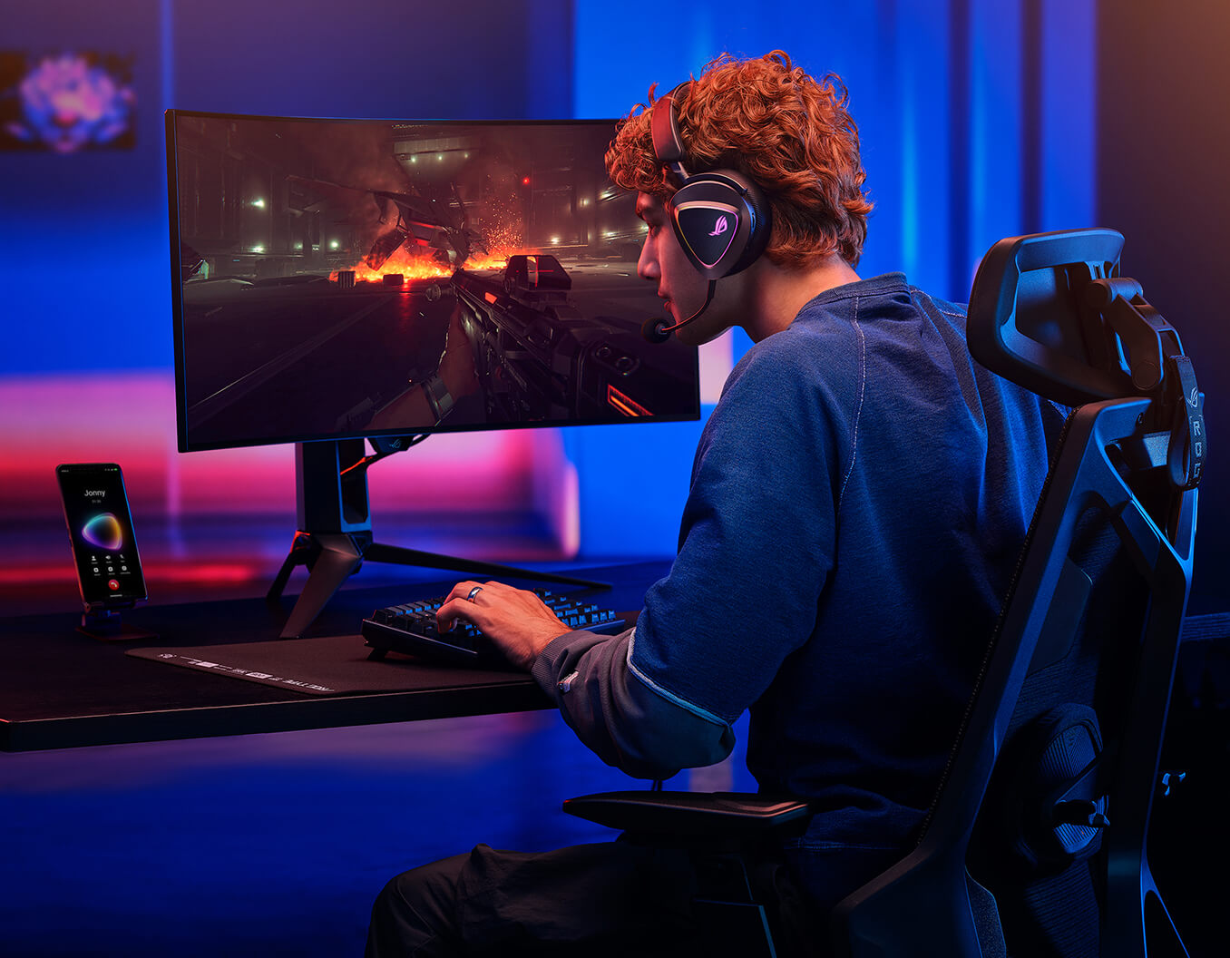 A man with curly hair sits at a ROG Destrier Core, wearing the ROG Delta II with a microphone and instense staring at the monitor. Viewing from the back, the ROG phone displaying the screen that he's during a phone call.