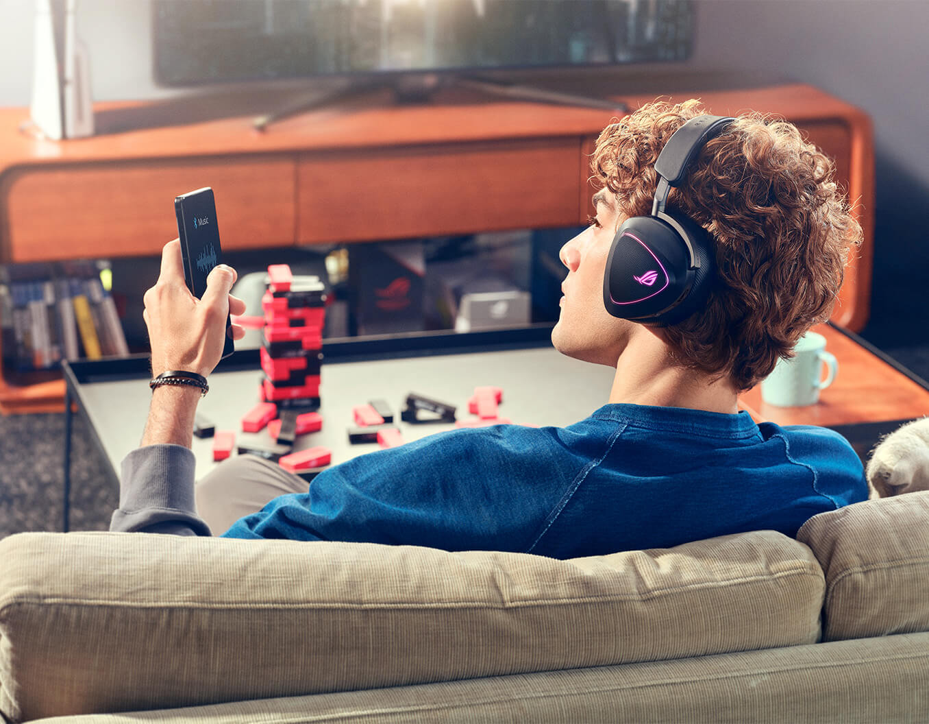 A man with curly hair wearing ROG Delta II sits on the sofa and holds a ROG phone on the left hand. The ROG phone displays the music listening app.