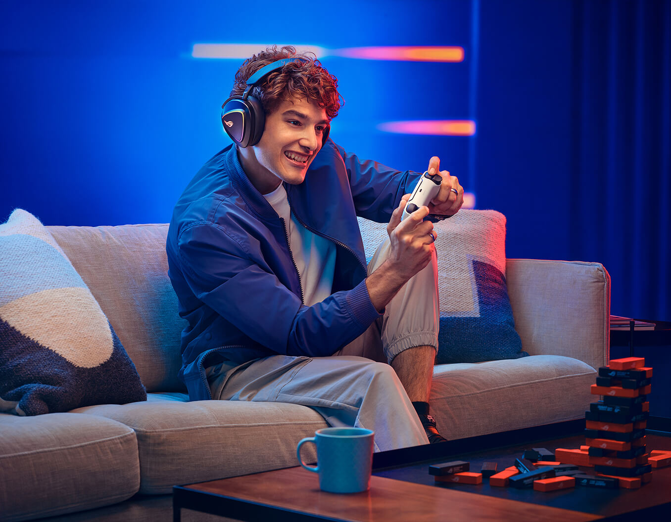 A man with curly hair wearing ROG Delta II sits on the sofa and holds a Playstation 5 controller.