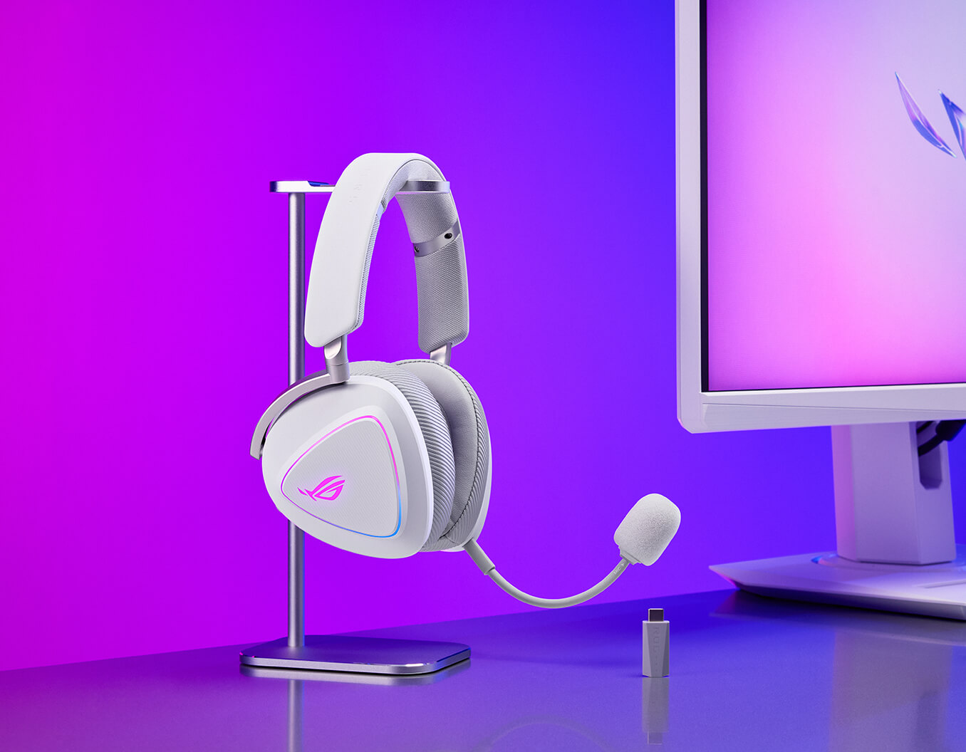 A gaming setup on a desk with a monitor. In front of the monitor are a ROG keyboard, a gaming mouse, and a ROG Carnyx. To the right of the monitor, a gaming headset with a glowing logo is placed on a sleek headset stand. The background features a gradient of purple and blue lighting, creating a vibrant and immersive atmosphere.
