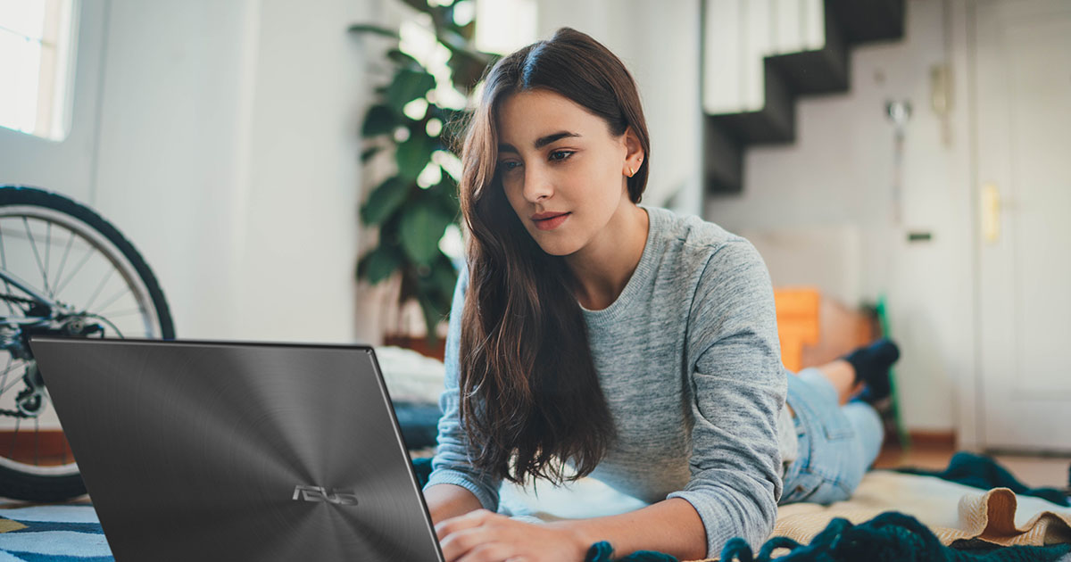 jeune femme habillée de façon décontractée, allongée sur le sol à la maison, utilisant l'ordinateur portable ASUS Zenbook 14X OLED