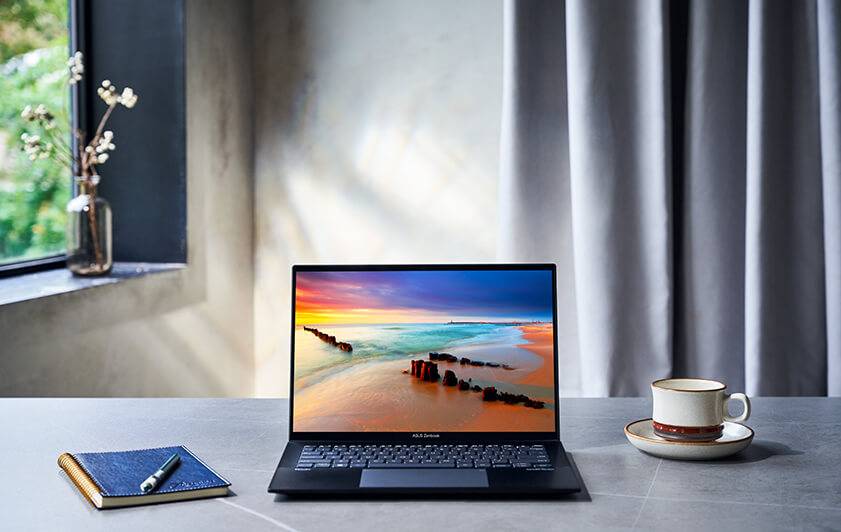 L'ordinateur portable ASUS Zenbook 14 OLED est posé sur la table, avec un ordinateur portable à gauche et une tasse à droite. La lumière du soleil vient de la fenêtre, ce qui éclaire toute la pièce.