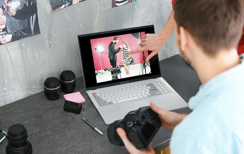  Un photographe tenant un appareil photo regarde la photo qu'il a prise du Vivobook Pro 16X OLED. Quelqu'un suggère la direction de la prise de vue pour le modèle.