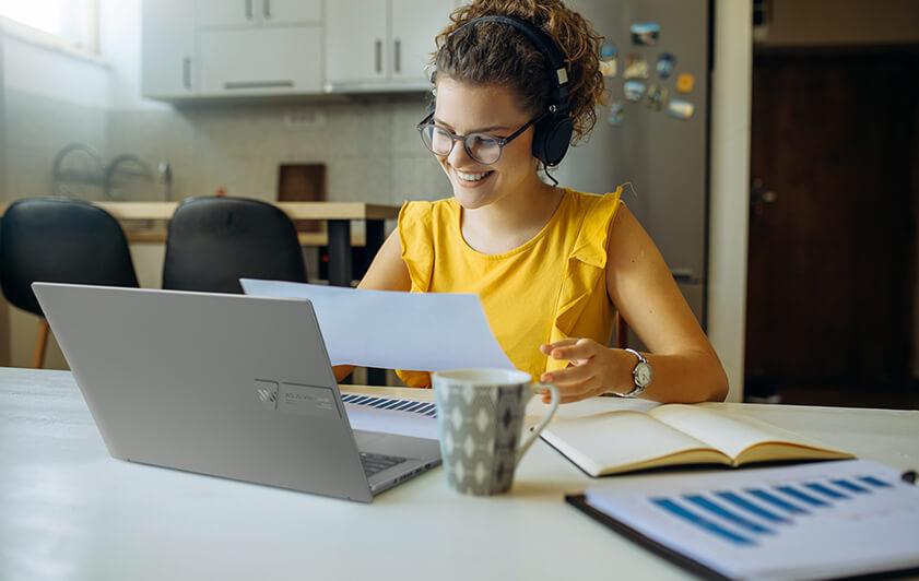 Une femme lit un document et écoute de la musique en même temps, avec le sourire. Le Vivobook S 14X OLED est devant elle.