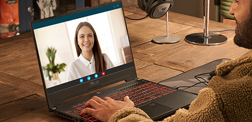 A person working at a wooden table with a laptop, with another person on screen for a video call.