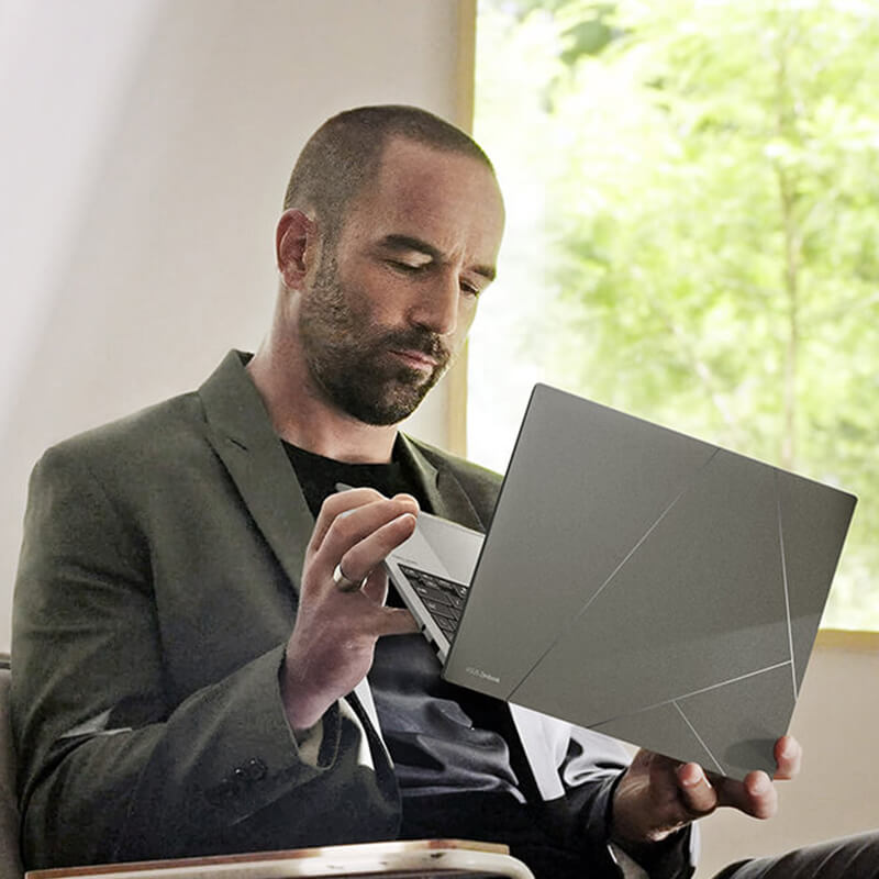 A man in smart casual attire is holding Zenbook S 13 OLED basalt grey version and inspecting its appearance. The background is beige with greenery on the right.