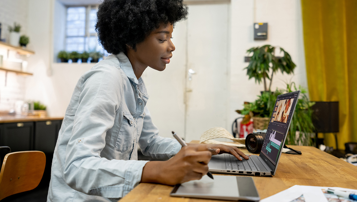 a young female content creator using pen tablet to edit videos on her ASUS Vivobook Pro 15 OLED creator laptop while sitting at a wooden table with a camera behind the laptop