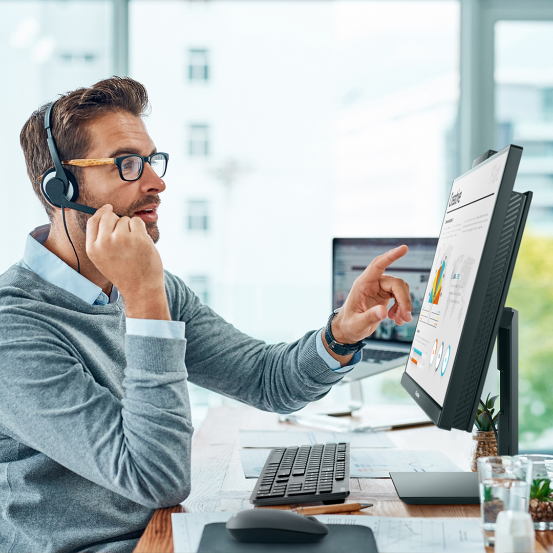 An employee is looking and pointing at his ASUS ExpertCenter AiO, talking on a call with headphones