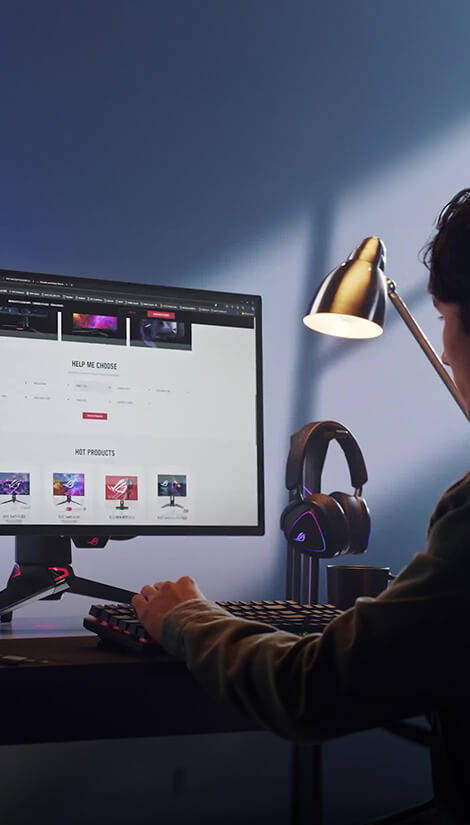 Muž píše na počítači, na stole má monitor, sluchátka, klávesnici, myš a malou lampu.