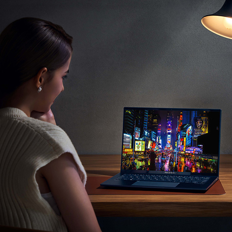 A woman watches a movie by a laptop in dark environment. The only light comes from the lamp on the table. The OLED screen displays a vivid city night view