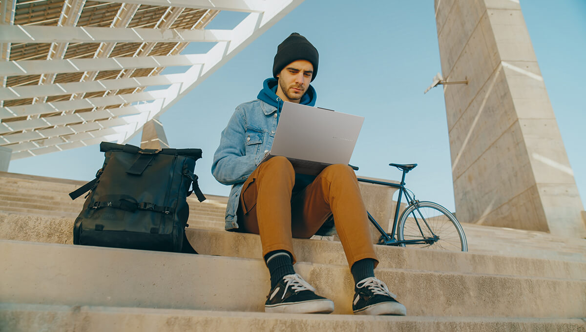 Young man sits on stairs outdoors with an ASUS Vivobook Pro 15 OLED laptop on his lap