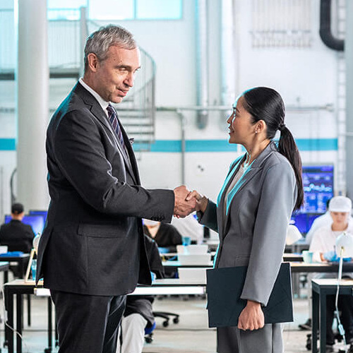 A man and a woman in suits are shaking hands in front of several rows of working people sitting at tables in lab coats.