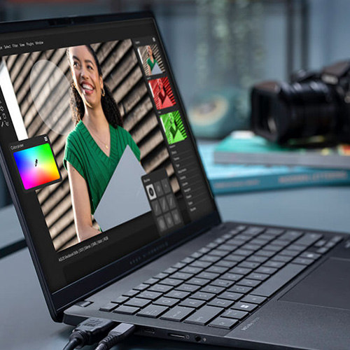 an ASUS Vivobook S 14 in neutral black on a desk, running an image editing application, with connected cables and a camera in the background.