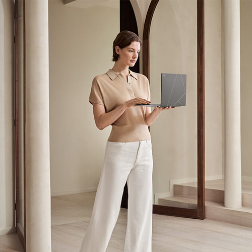 A young woman standing in a light-filled living room holding an open Zenbook S 14 laptop in her hands