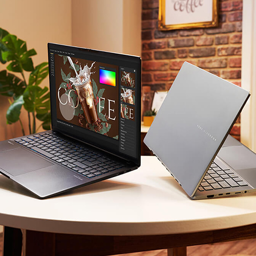 two open ASUS Vivobook S14 laptops in Neutral Black and Cool Silver colors lying on a table in a warm-colored, well-lit room