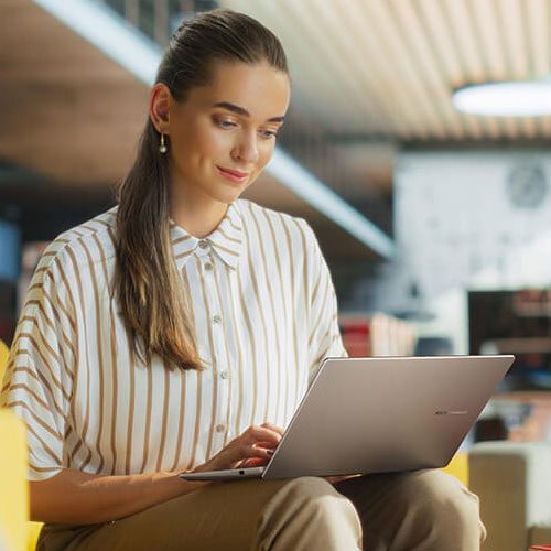 A woman is using a silver ASUS Vivobook on a yellow sofa in a lounge setting.