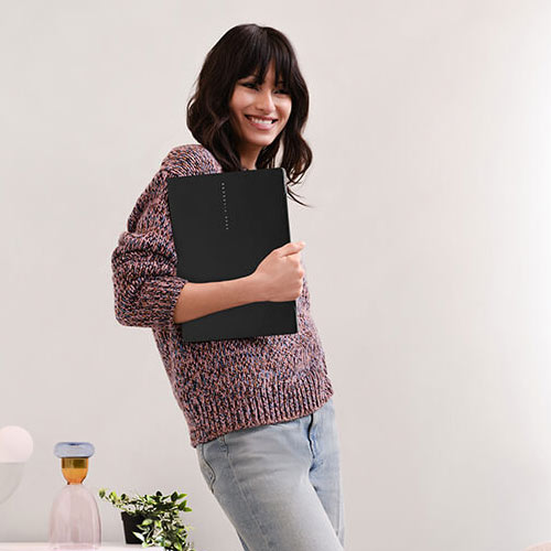 a young, smiling woman standing in a colorful living room, holding an ASUS Vivobook S14 laptop in her right hand