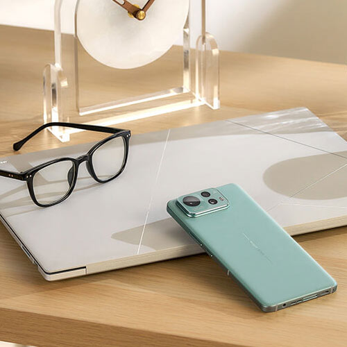 a green Zenfone 12 Ultra leaning against an ASUS Zenbook laptop, placed on a brightly-colored wooden desk, with everyday objects lying around it