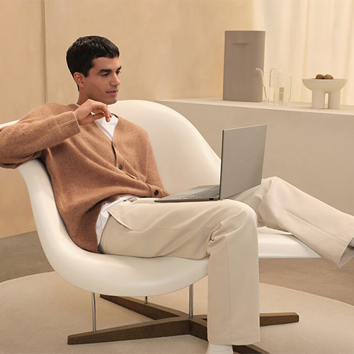 A man sits in a comfortable, laid-back position on a white designer armchair with an ASUS Zenbook A14 laptop on his lap, looking at its screen