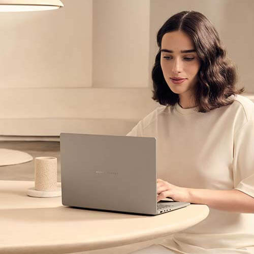 A woman in a beige outfit is sitting at a table using ASUS Zenbook A14.