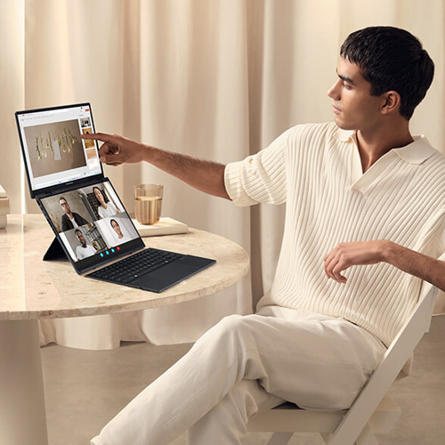 a young, brightly-dressed man touches the top screen of an ASUS Zenbook DUO laptop while displaying a Powerpoint presentation in a team meeting on the bottom screen