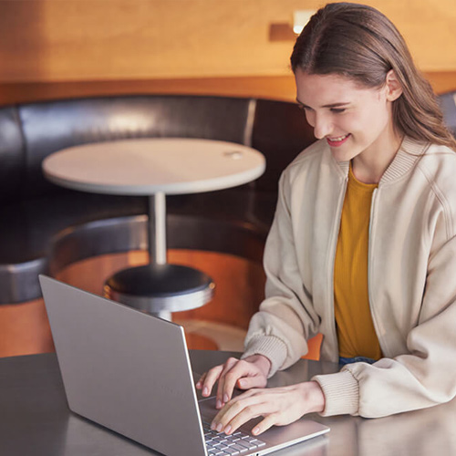 A woman smiles as she sits in a café typing on her ASUS Vivobook S 16 laptop