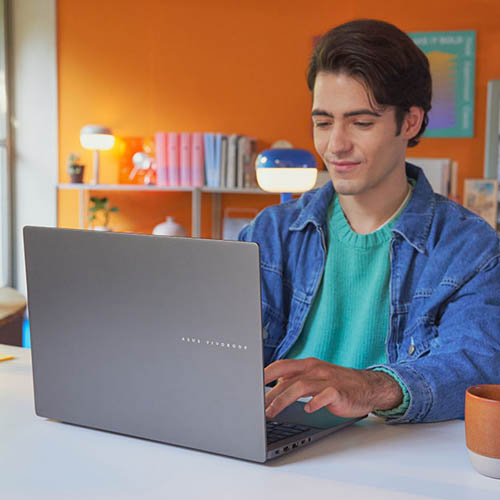 genç bir adam, parlak renkli bir odada oturan ASUS Vivobook S16 dizüstü bilgisayarını kullanırken gülümsüyor