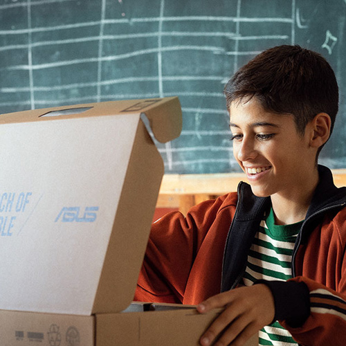 Улыбающийся школьник открывает коробку с ноутбуком ASUS, сидя за партой на фоне классной доски 