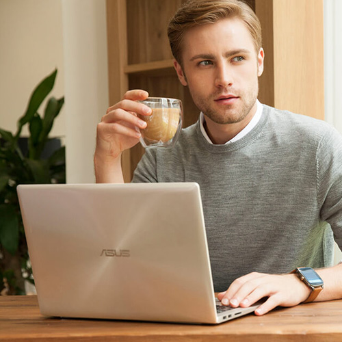 Мужчина кладёт правую руку на ноутбук ASUS, лежащий на столе. В другой руке он держит чашку кофе и глядит на улицу.