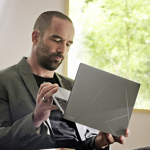 Мужчина, одетый в стиле смарт-кэжуал, держит в руках ноутбук Zenbook S 13 OLED в оформлении Basalt Grey. Фон – бежевая комната с деревьями за окном.