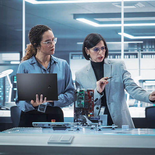 Two women are in a laboratory discussing and researching. The one on the left is wearing a blue shirt and holding an ASUS laptop. The one on the right is in a grey blazer and gesturing to the right in mid-conversation.