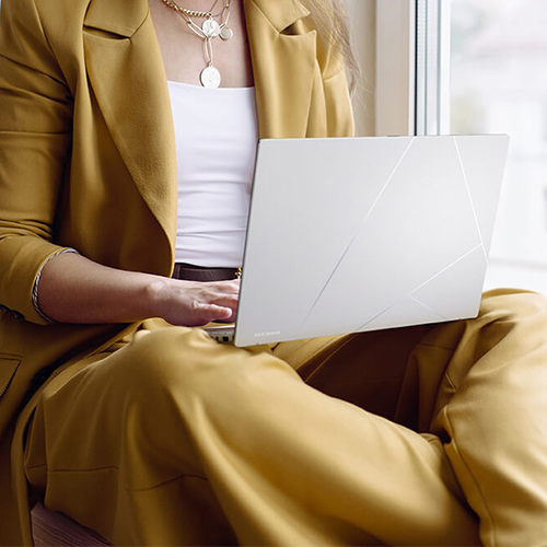 A woman in a yellow suit sits by a window with a Zenbook 14 OLED on her lap.